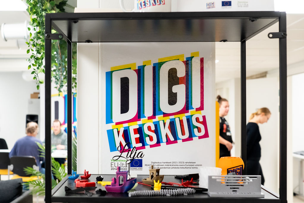Digikeskuksen 3D-tulostusesimerkkejä hyllyllä ja taustalla Digikeskuksen logo